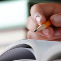 Auf einem aufgeschlagenen, dicken Heft setzt eine Hand in Großaufnahme mit einem gelben Bleistift zum Schreiben an.